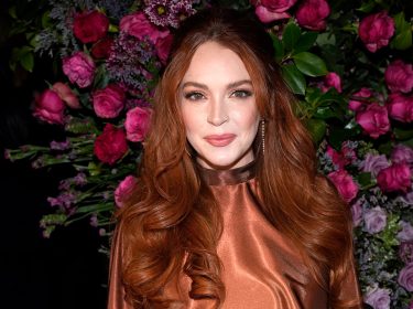 Auf dem Eventfoto ist Lindsay Lohan mit offenen, gewellten roten Haaren und einem schimmernden, bronzefarbenen Kleid zu sehen. Sie steht vor einer üppigen Blumenwand aus violetten und rosafarbenen Rosen und schaut direkt in die Kamera. Das Bild wird im Zusammenhang mit der möglicherweise geplanten Serie Count My Lies verwendet