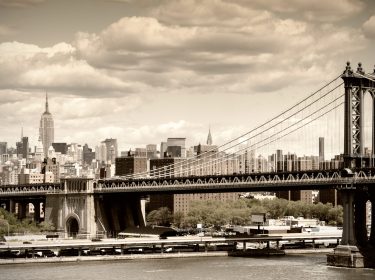Auf diesem Symbolbild zur Zeit der Unschuld-Serie ist die Manhattan Bridge im Vordergrund zu sehen, während im Hintergrund die Skyline von New York mit dem Empire State Building zu sehen ist. Das Bild ist in sepiafarbenem Ton gehalten und vermittelt eine nostalgische, historische Atmosphäre, die an das New York der 1920er Jahre erinnert.