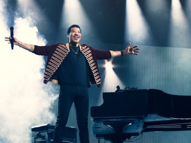 Im Artikel zu den Lionel Richie Tickets zeigt dieses Bild den Künstler in einem eindrucksvollen Moment auf der Bühne: Mit ausgebreiteten Armen steht Lionel Richie neben einem schwarzen Konzertflügel, umgeben von dichter Nebelstimmung und dramatischer Beleuchtung.