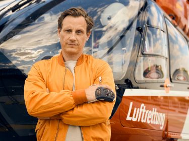 Auf dem Bild zu den ZDF-Serien Empfehlungen ist Schauspieler Patrick Kalupa als Dr. Moritz Neiss zu sehen. Er steht mit verschränkten Armen und ernstem Blick vor einem Rettungshubschrauber mit der Aufschrift „Luftrettung“. Kalupa trägt eine orangefarbene Fliegerjacke, was seine Rolle als Notarzt unterstreicht. Der Himmel spiegelt sich dramatisch in der Cockpit-Scheibe.