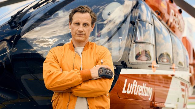 Auf dem Bild zu den ZDF-Serien Empfehlungen ist Schauspieler Patrick Kalupa als Dr. Moritz Neiss zu sehen. Er steht mit verschränkten Armen und ernstem Blick vor einem Rettungshubschrauber mit der Aufschrift „Luftrettung“. Kalupa trägt eine orangefarbene Fliegerjacke, was seine Rolle als Notarzt unterstreicht. Der Himmel spiegelt sich dramatisch in der Cockpit-Scheibe.