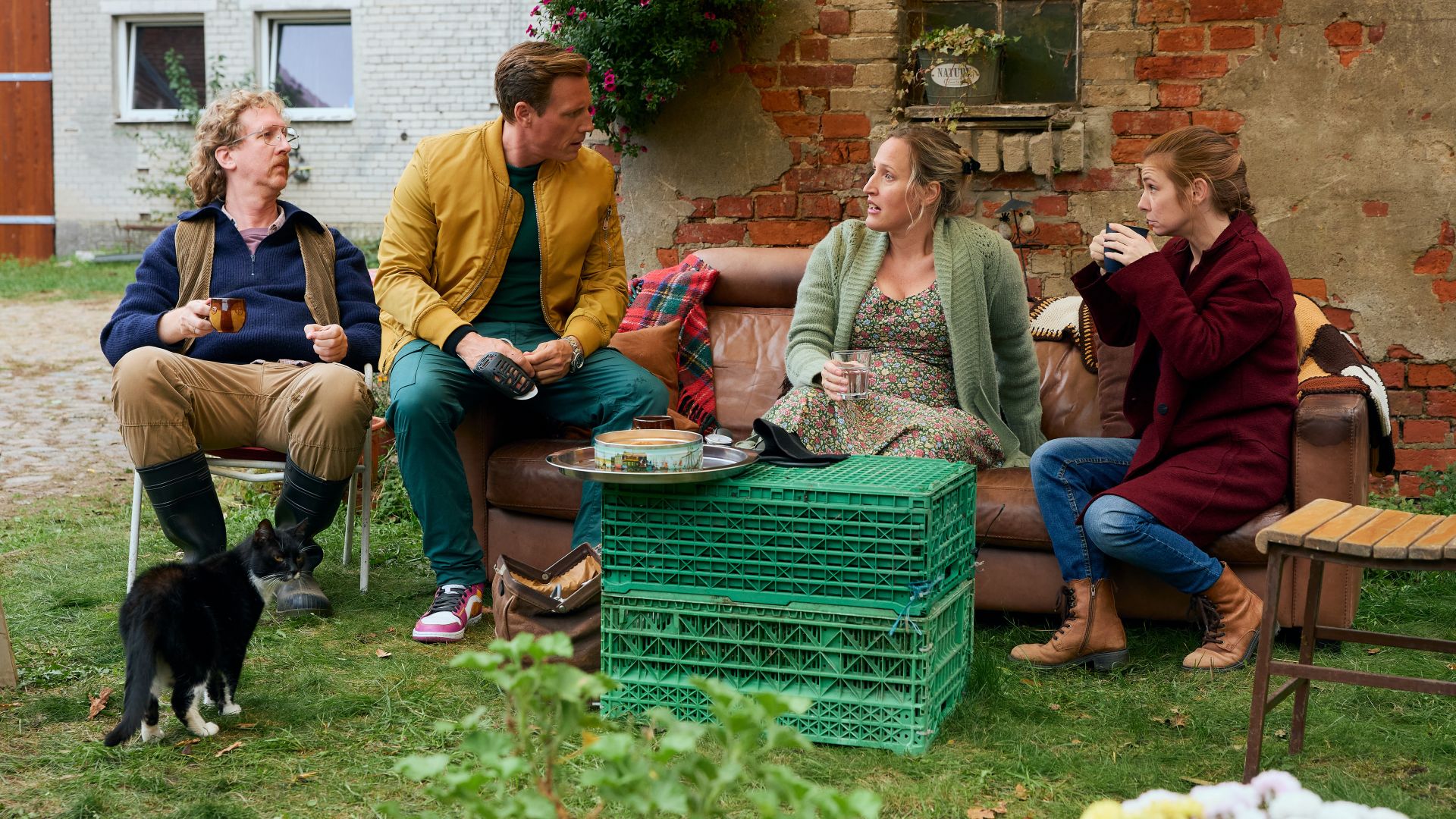 Uke Bosse, Patrick Kalupa, Luise Schnittert und Josefine Preuß sitzen in einer Szene von Dr. Nice während einer Teepause im Garten eines Bauernhofes.