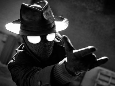 In dieser Szene aus der Spider-Noir Serie ist Spider-Man-Noir, gespielt von Nicolas Cage, in einem stilisierten Schwarz-Weiß-Look zu sehen. Er trägt einen Fedora-Hut, eine Gesichtsmaske und einen langen Mantel.