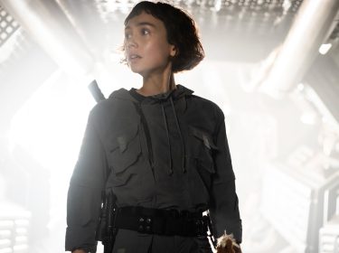 In dieser Szene aus der Alien: Earth Serie steht Wendy, gespielt von Sydney Chandler, in einem futuristischen Raumschiffkorridor. Sie trägt einen dunklen Kampfanzug mit taktischem Gürtel, wirkt angespannt und blickt nach links.