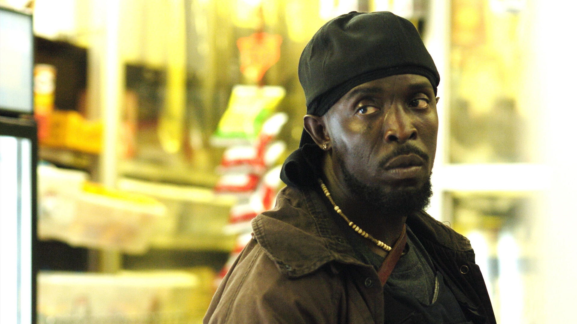 Auf diesem Bild zur „The Wire“-Serie ist Omar Little zu sehen, gespielt von Michael Kenneth Williams. Er steht in einem kleinen Laden, trägt eine dunkle Jacke, ein Bandana und eine Perlenkette. Sein ernster, misstrauischer Blick unterstreicht die angespannte Stimmung der Szene, typisch für die düstere Atmosphäre der The Wire-Serie.