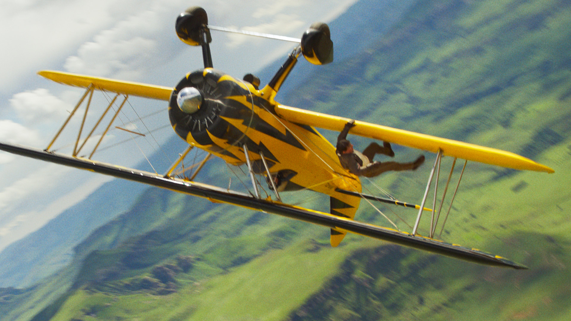 Im Bild zur Mission: Impossible 8 Kritik ist Tom Cruise als Ethan Hunt in einer waghalsigen Actionszene zu sehen. Er hängt kopfüber an einem gelb-schwarzen Doppeldeckerflugzeug, das sich in rasanter Schräglage über eine grüne Hügellandschaft bewegt. Die Szene vermittelt Spannung, Risiko und den für die Filmreihe typischen Adrenalinrausch.