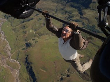 Im Bild zur Mission: Impossible 8 Kritik hängt Tom Cruise als Ethan Hunt in schwindelerregender Höhe an einem Helikopterfahrwerk über einer zerklüfteten Landschaft. Mit Schutzbrille und konzentriertem Gesichtsausdruck stemmt er sich gegen den Wind. Die Szene betont erneut die kompromisslose Action und den körperlichen Einsatz, den die Reihe berühmt gemacht hat.