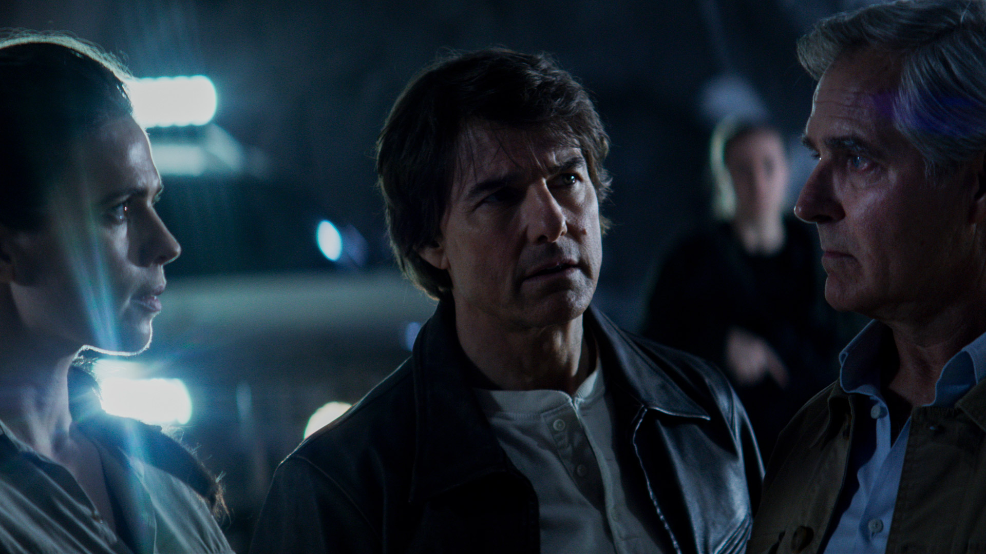 Im Bild zur Mission: Impossible 8 Kritik stehen Hayley Atwell als Grace, Tom Cruise als Ethan Hunt und Henry Czerny als Kittridge angespannt beieinander. Die Szene spielt in einer düsteren Umgebung mit starker Gegenlichtbeleuchtung, was die Dramatik und Ernsthaftigkeit ihrer Besprechung unterstreicht. Ihre Blicke sind konzentriert, die Stimmung wirkt angespannt und entscheidend.