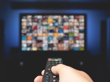 Auf diesem Symbolbild zur Frage „Was kostet Sky 2025“ ist eine Hand mit Fernbedienung zu sehen, die auf einen unscharfen Fernseher mit zahlreichen Streaming-Inhalten gerichtet ist. Das Bild symbolisiert die Vielfalt an Programmen und Paketen, die Sky 2025 bietet, und eignet sich ideal zur Illustration von Kostenübersichten zu Abos und Streaming-Angeboten.