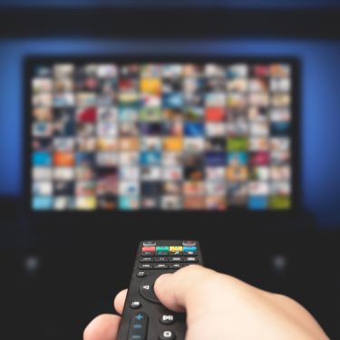 Auf diesem Symbolbild zur Frage „Was kostet Sky 2025“ ist eine Hand mit Fernbedienung zu sehen, die auf einen unscharfen Fernseher mit zahlreichen Streaming-Inhalten gerichtet ist. Das Bild symbolisiert die Vielfalt an Programmen und Paketen, die Sky 2025 bietet, und eignet sich ideal zur Illustration von Kostenübersichten zu Abos und Streaming-Angeboten.