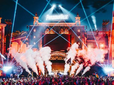 Bei diesem Bild zum Thema parookaville tickets handelt es sich um eine energiegeladene Liveszene des Parookaville-Festivals bei Nacht. Die große Hauptbühne ist festlich beleuchtet, mit leuchtenden Lasern, Rauchfontänen und einer Menschenmenge voller feiernder Besucher im Vordergrund.
