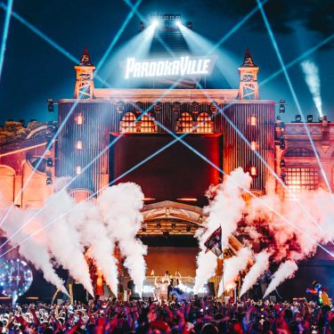 Bei diesem Bild zum Thema parookaville tickets handelt es sich um eine energiegeladene Liveszene des Parookaville-Festivals bei Nacht. Die große Hauptbühne ist festlich beleuchtet, mit leuchtenden Lasern, Rauchfontänen und einer Menschenmenge voller feiernder Besucher im Vordergrund.