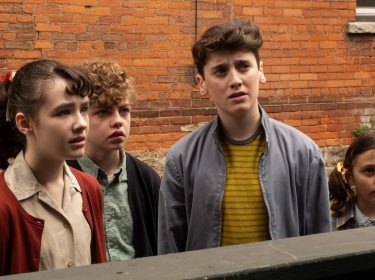 Welcome to Derry zeigt in dieser Szene vier Kinder, die mit ernsten und teils verunsicherten Blicken in dieselbe Richtung schauen. Die Gruppe steht vor einer Backsteinwand, ihre Kleidung wirkt retro und deutet auf eine frühere Zeitperiode hin.