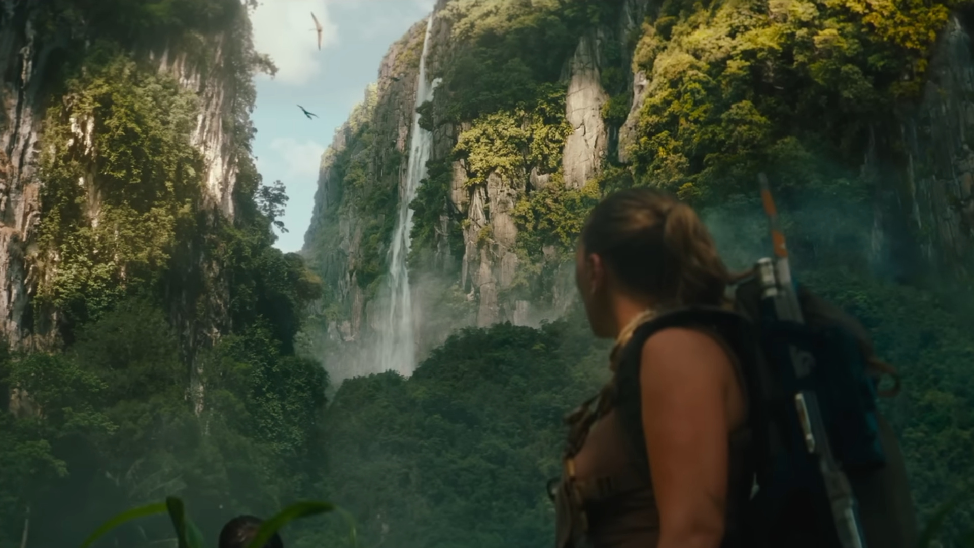 Bild aus Jurassic World: Die Wiedergeburt - Eine Abenteurerin mit Rucksack blickt auf ein riesiges Tal mit Wasserfällen und fliegenden Dinosauriern.