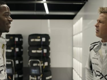 Bild aus F1: Zwei Rennfahrer in weißer Teamkleidung stehen in der Boxengasse und unterhalten sich lachend vor einem Stapel Rennreifen.