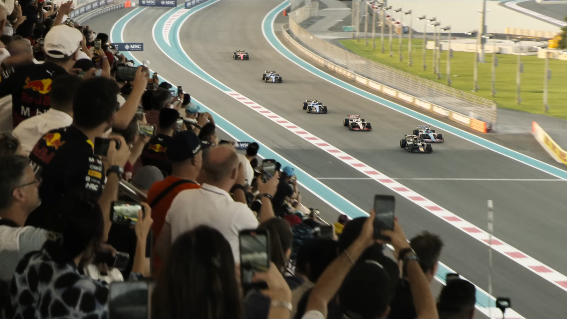 Bild aus dem Film F1: Formel-1-Autos rasen auf der Strecke, während jubelnde Zuschauer in der Tribüne das Rennen mit ihren Smartphones festhalten.
