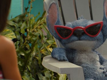 Auf dem Bild in dem Artikel zum Lilo & Stitch-Vergleich ist Stitch in der Live-Action-Version von 2025 in einer entspannten Szene zu sehen. Er sitzt lässig auf einem weißen Gartenstuhl, trägt eine rote Retro-Sonnenbrille und blickt cool zur Seite. Neben ihm ist unscharf Lilo zu erkennen.
