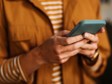 Auf dem Bild zur Nutzung von Meta AI bei WhatsApp ist eine Person zu sehen, die in einem Alltagsszenario ein Smartphone in der Hand hält. Die Person trägt eine gelb-braune Jacke über einem gestreiften Shirt und schaut konzentriert auf das Display. Die Szene vermittelt einen modernen und fokussierten Moment digitaler Kommunikation.