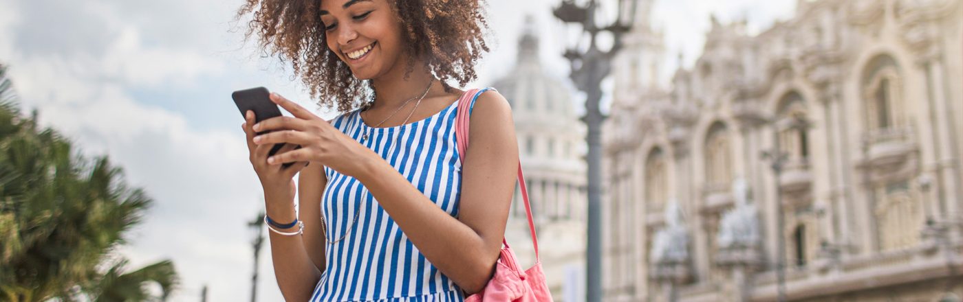 Eine junge Frau steht im Sommerkleid mit blau-weißen Streifen lachend auf einer belebten Straße und schaut auf ihr Smartphone. Im Hintergrund sind prunkvolle historische Gebäude und Palmen zu sehen. Die Szene vermittelt Leichtigkeit und Entdeckerfreude – ideal passend zum Thema Roaming und mobiles Internet auf Reisen.