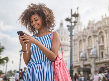 Eine junge Frau steht im Sommerkleid mit blau-weißen Streifen lachend auf einer belebten Straße und schaut auf ihr Smartphone. Im Hintergrund sind prunkvolle historische Gebäude und Palmen zu sehen. Die Szene vermittelt Leichtigkeit und Entdeckerfreude – ideal passend zum Thema Roaming und mobiles Internet auf Reisen.