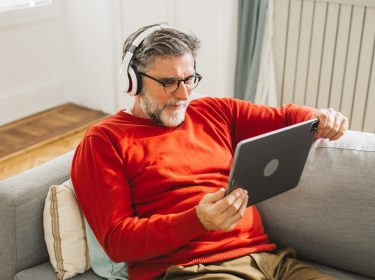 Ein älterer Mann mit grauem Haar und Bart sitzt entspannt auf einem hellgrauen Sofa und schaut mit Kopfhörern ein Video auf einem Tablet. Er trägt einen roten Pullover und wirkt konzentriert und zufrieden. Dieses Bild symbolisiert bequemes Streaming von zuhause und passt thematisch zur Darstellung der ARD Plus Kosten.