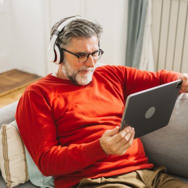 Ein älterer Mann mit grauem Haar und Bart sitzt entspannt auf einem hellgrauen Sofa und schaut mit Kopfhörern ein Video auf einem Tablet. Er trägt einen roten Pullover und wirkt konzentriert und zufrieden. Dieses Bild symbolisiert bequemes Streaming von zuhause und passt thematisch zur Darstellung der ARD Plus Kosten.