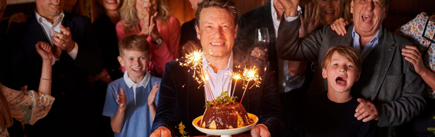 In dem Artikel zu den kostenlosen Jamie Oliver Folgen zu seinem 50. Geburtstag ist Jamie Oliver im Mittelpunkt einer festlichen Szene zu sehen. Er lächelt breit, hält einen Teller mit Schokopudding und Wunderkerzen in der Hand, während hinter ihm eine Gruppe aus Kindern und Erwachsenen applaudiert und feiert.