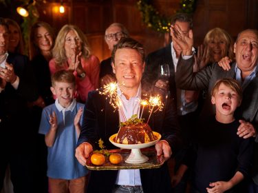 In dem Artikel zu den kostenlosen Jamie Oliver Folgen zu seinem 50. Geburtstag ist Jamie Oliver im Mittelpunkt einer festlichen Szene zu sehen. Er lächelt breit, hält einen Teller mit Schokopudding und Wunderkerzen in der Hand, während hinter ihm eine Gruppe aus Kindern und Erwachsenen applaudiert und feiert.