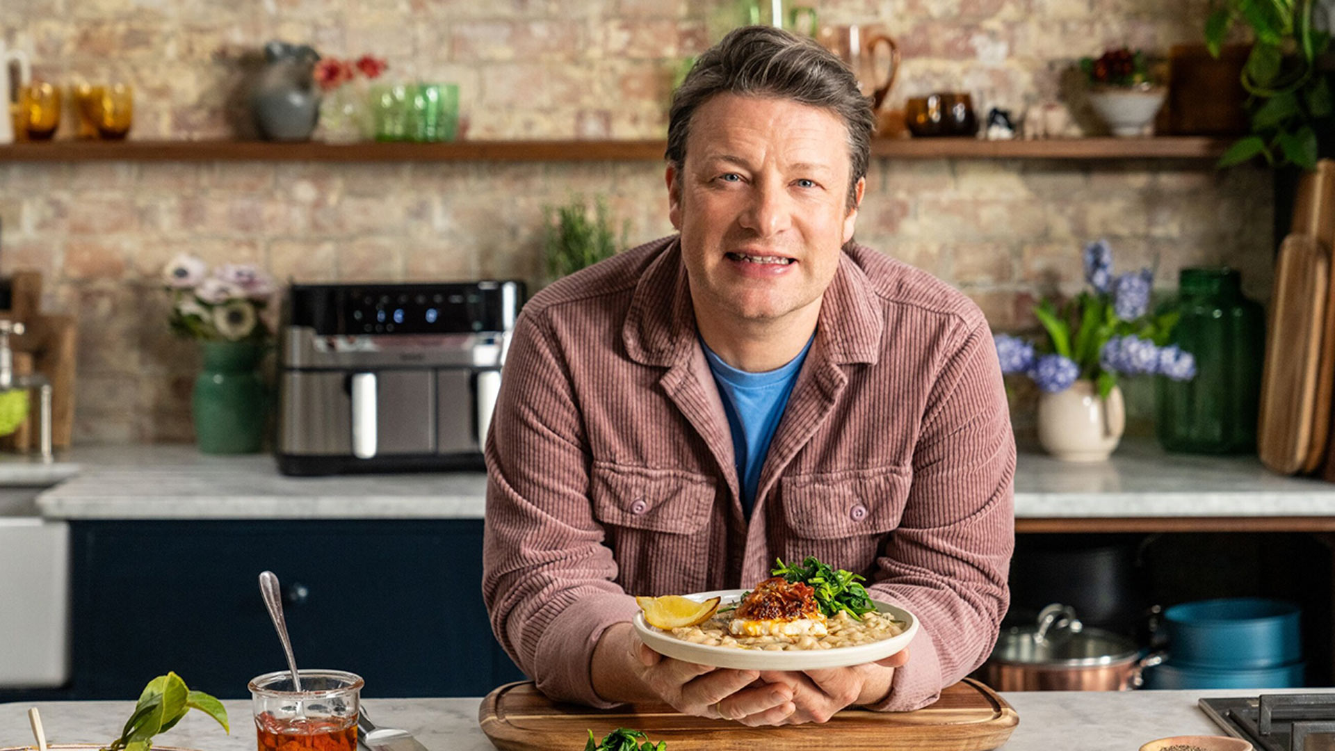 In dem Artikel zu den kostenlosen Jamie Oliver Folgen zu seinem 50. Geburtstag ist Jamie Oliver in einer gemütlichen Studioküche zu sehen. Er trägt eine rosa Cordjacke, blickt freundlich in die Kamera und präsentiert ein liebevoll angerichtetes Tellergericht mit Fisch, Bohnen und Gemüse.
