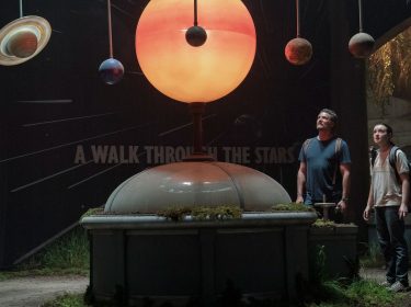 Auf dem Bild zur The Last of Us Staffel 3 sind Pedro Pascal als Joel und Bella Ramsey als Ellie zu sehen. Sie stehen in einem verlassenen Museum unter einem beleuchteten Modell des Sonnensystems mit Planeten. Beide blicken fasziniert nach oben, die Szene wirkt ruhig und andächtig, fast magisch – ein seltener Moment der Hoffnung in einer postapokalyptischen Welt.