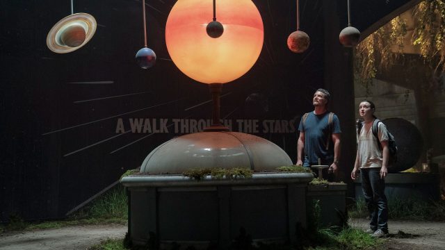 Auf dem Bild zur The Last of Us Staffel 3 sind Pedro Pascal als Joel und Bella Ramsey als Ellie zu sehen. Sie stehen in einem verlassenen Museum unter einem beleuchteten Modell des Sonnensystems mit Planeten. Beide blicken fasziniert nach oben, die Szene wirkt ruhig und andächtig, fast magisch – ein seltener Moment der Hoffnung in einer postapokalyptischen Welt.