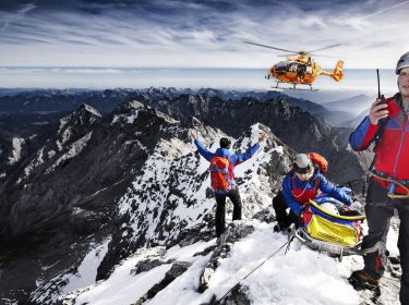 Drei Mitarbeiter:innen der Bergwacht, die sich mit einer geretteten Person auf einem Berggipfel befinden, weisen einen anfliegenden Hubschrauber ein.