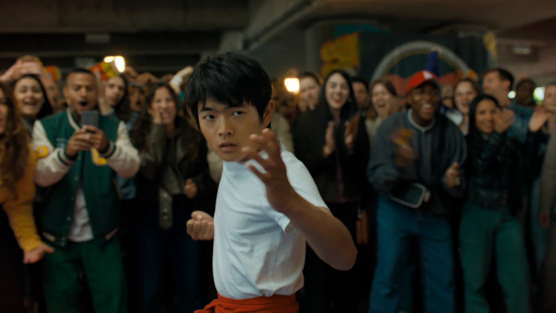 Ben Wang steht in dieser Szene aus Karate Kid: Legends kampfbereit in einem weißen Shirt mit rotem Gürtel in einer Menschenmenge. Mit entschlossenem Blick und fokussierter Haltung signalisiert er Bereitschaft und Mut. Hinter ihm jubelt eine diverse Gruppe junger Zuschauer – ein emotionaler Moment für die Karate Kid: Legends Kritik.