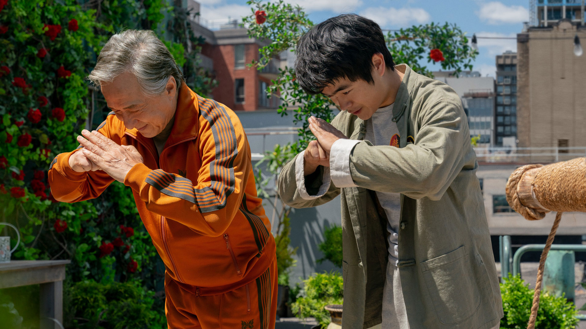 Im Bild aus Karate Kid: Legends verbeugen sich Jackie Chan und Ben Wang respektvoll voreinander in einem sonnigen Dachgarten. Jackie Chan trägt einen auffälligen orangefarbenen Trainingsanzug, während Ben Wang eine grüne Jacke mit Emblem trägt. Die Szene strahlt Ruhe, Respekt und Verbundenheit aus – passend zur Karate Kid: Legends Kritik