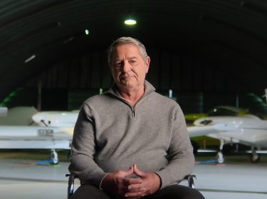 Produktionsstandbild aus der Netflix-Doku „Air Cocaine“: Ein älterer Mann sitzt in einem Flugzeughangar, im Hintergrund stehen zwei kleine Maschinen.