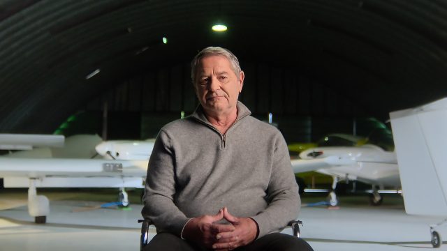 Produktionsstandbild aus der Netflix-Doku „Air Cocaine“: Ein älterer Mann sitzt in einem Flugzeughangar, im Hintergrund stehen zwei kleine Maschinen.