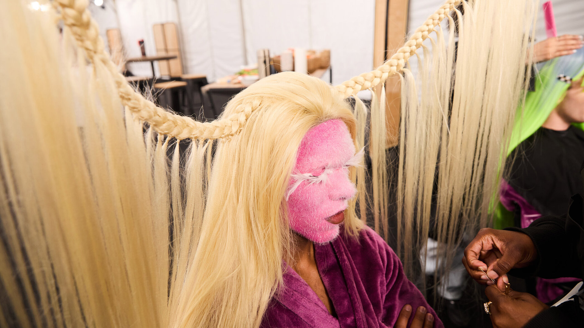 Auf dem Bild aus GNTM 2025 ist Eliob (29) während seiner Verwandlung zum haarigen Alien zu sehen. Sein Gesicht ist vollständig pink beflockt, mit flauschigen Wimpern, und sein platinblondes Haar ist seitlich zu einem auffälligen, waagerechten Zopf gespannt. Die Szene wirkt surreal und kreativ – typisch für ein GNTM-Umstyling.