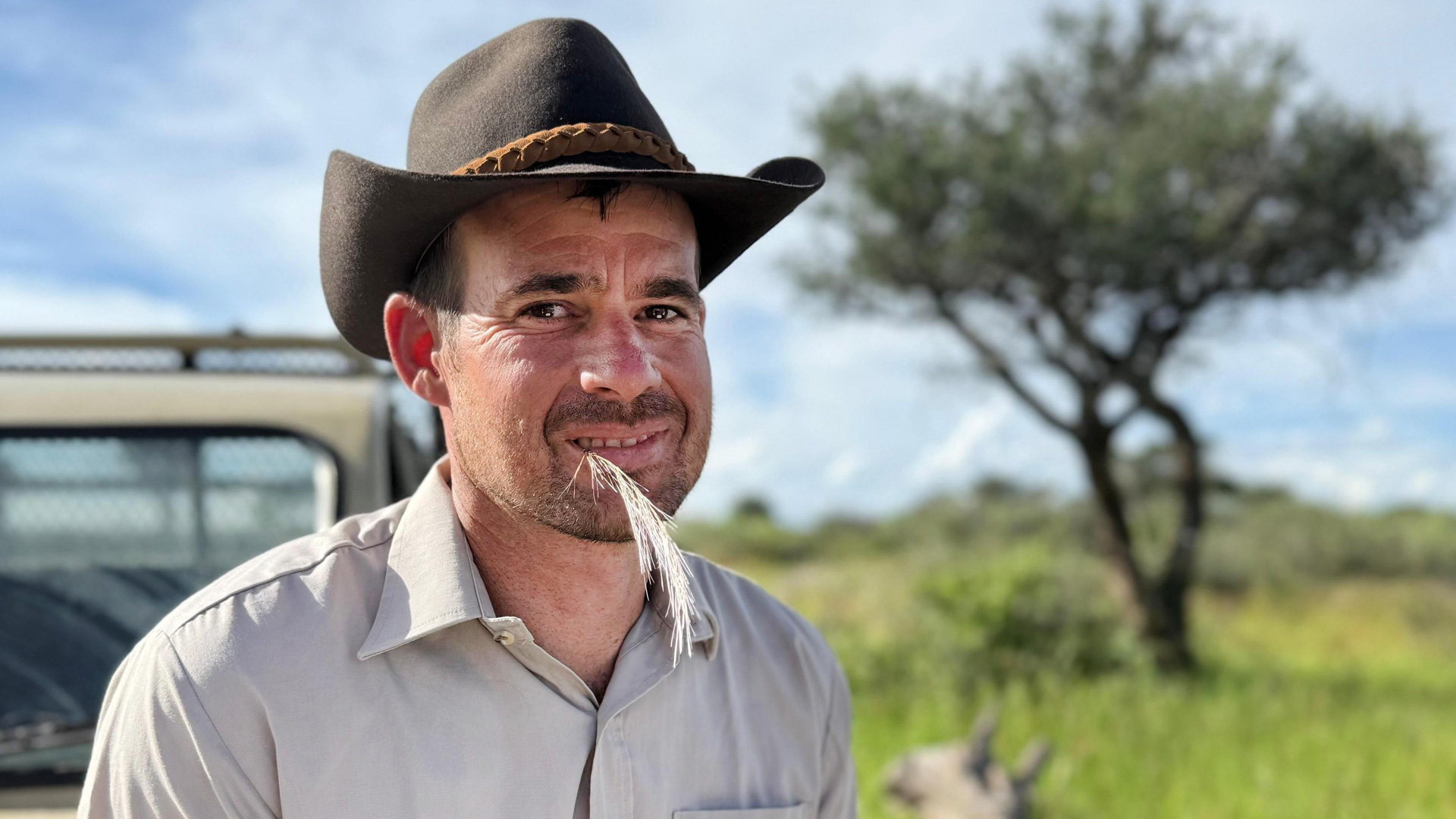Marco, Kandidat der RTL-Sendung „Bauer sucht Frau international Staffel 7“, steht im Halbschatten eines Baums in der weiten Savannenlandschaft Namibias. Er trägt ein helles Hemd und einen braunen Cowboyhut und hat ein trockenes Gras zwischen den Lippen. Im Hintergrund sind ein Geländewagen und ein einzelner Akazienbaum zu sehen – die Szene wirkt ruhig und naturverbunden.
