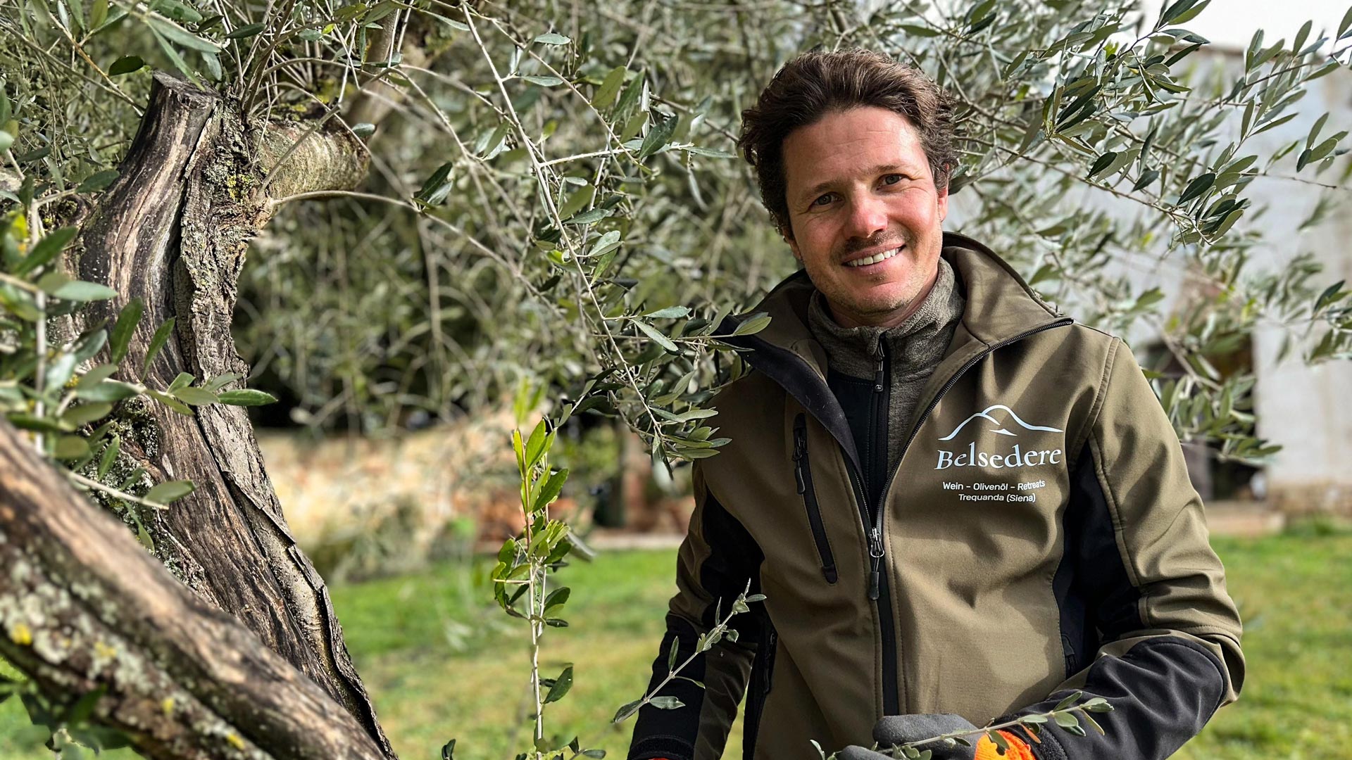 Martin, Teilnehmer der RTL-Sendung „Bauer sucht Frau international Staffel 7“, steht zwischen den Zweigen eines Olivenbaums auf seinem Hof in der Toskana. Er trägt eine Jacke mit der Aufschrift „Belsedere“ und lächelt freundlich. Die Szene wirkt ruhig und naturverbunden, mit grünem Rasen und traditioneller Architektur im Hintergrund.
