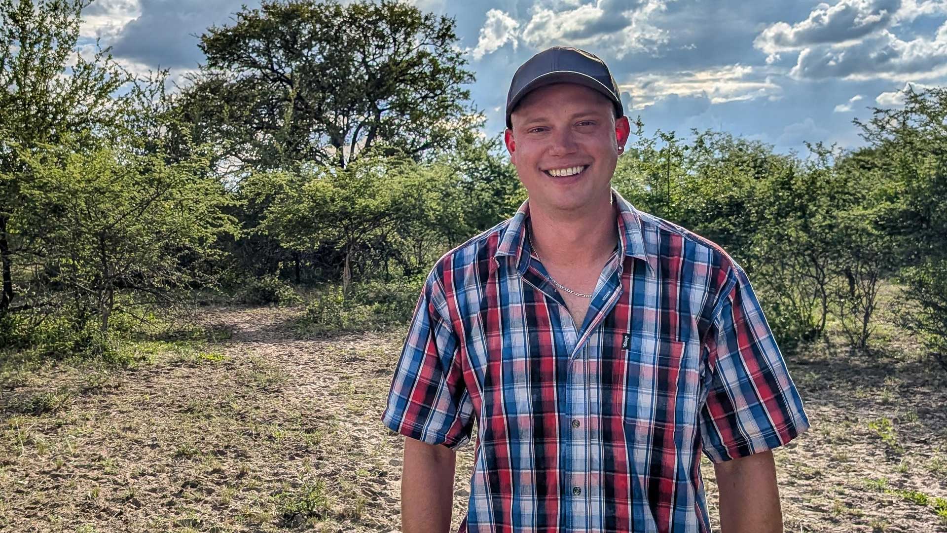 Björn, Kandidat der RTL-Sendung „Bauer sucht Frau international Staffel 7“, steht lachend auf trockenem Boden in der savannenartigen Landschaft Namibias. Er trägt ein kariertes Hemd in Blau-Rot und eine dunkle Cap. Die Sonne scheint durch eine aufgelockerte Wolkendecke, während Büsche und Bäume im Hintergrund das ländliche Setting betonen.
