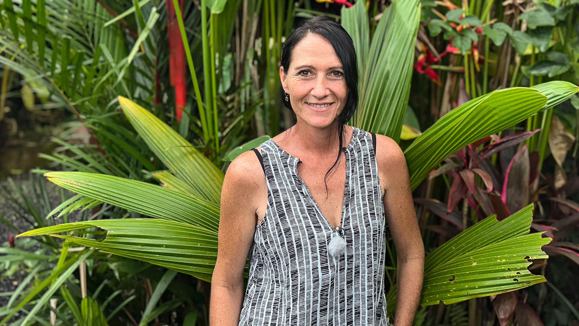 Carmen, eine Teilnehmerin der RTL-Sendung „Bauer sucht Frau international Staffel 7“, steht lächelnd vor tropischen Pflanzen auf Bali. Sie trägt ein ärmelloses Oberteil mit schwarz-weißem Muster. Die Kulisse mit großen, grünen Blättern und exotischen Farben unterstreicht die tropische Stimmung des Settings und betont Carmens offene Ausstrahlung.