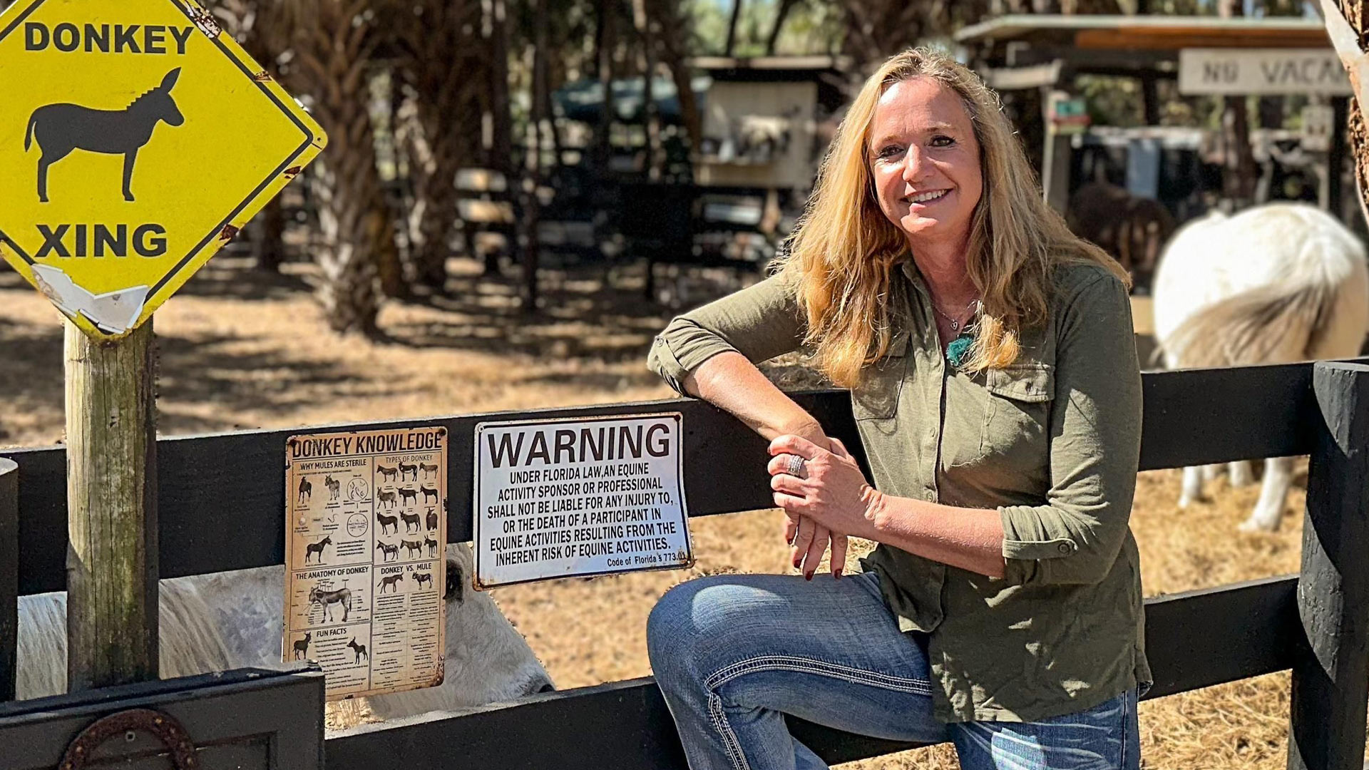 Iris, Kandidatin der RTL-Sendung „Bauer sucht Frau international Staffel 7“, lehnt entspannt an einem Zaun auf ihrer Farm in Florida. Im Hintergrund sind ein Esel, ein Pferd und mehrere Hinweisschilder zu sehen, darunter ein gelbes „Donkey Xing“-Schild. Sie trägt Jeans und ein grünes Hemd und lächelt freundlich in die Kamera bei sonnigem Wetter.
