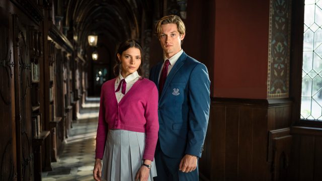 Auf dem Bild zur "Maxton Hall" Staffel 3 sind Harriet Herbig-Matten als Ruby Bell und Damian Hardung als James Beaufort in ihren Schuluniformen zu sehen. Ruby trägt eine pinke Strickjacke und einen grauen Faltenrock, James einen blauen Blazer mit Wappen. Beide stehen in einem historischen Schulflur mit dunklem Holz und wirken ernst und fokussiert.