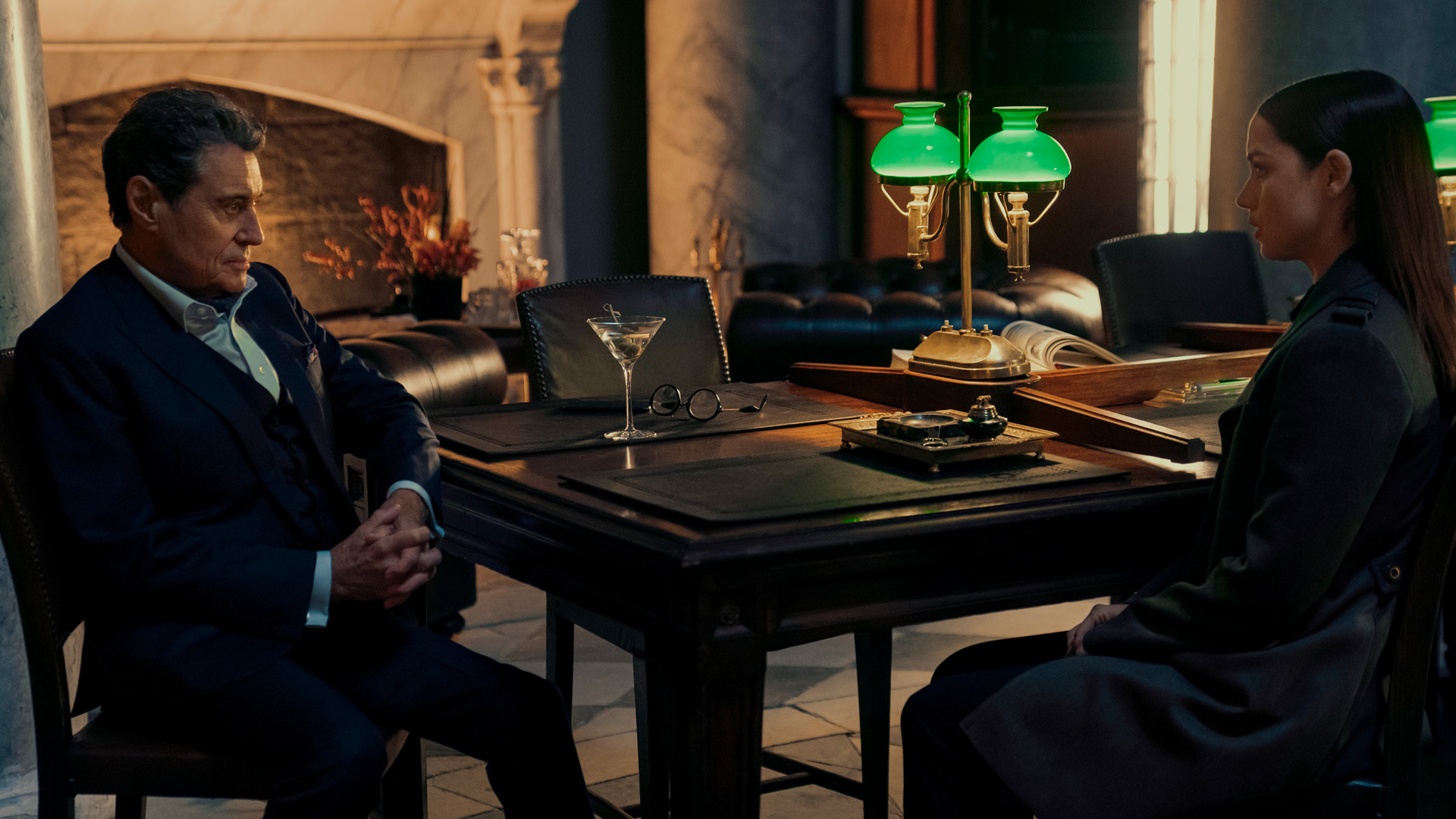 Im Bild zur John Wick Ballerina Kritik sitzen Ian McShane als Winston Scott und Ana de Armas als Eve sich in einem eleganten, dunklen Raum gegenüber. Winston trägt einen Anzug und hat die Hände gefaltet, während Eve ernst zuhört. Auf dem Tisch stehen eine grüne Bankerlampe, ein Martini-Glas und eine Brille, was die gehobene Atmosphäre unterstreicht.