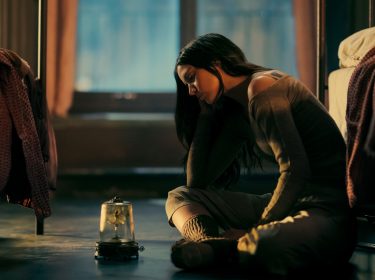 In dieser melancholischen Szene zur John Wick Ballerina Kritik sitzt Ana de Armas als Eve gedankenverloren auf dem Boden eines Schlafsaals zwischen zwei Betten. Sie trägt gemütliche Kleidung und blickt traurig auf eine Spieluhr mit einer tanzenden Figur unter einer Glaskuppel. Das warme Licht unterstreicht die emotionale Tiefe des Moments.