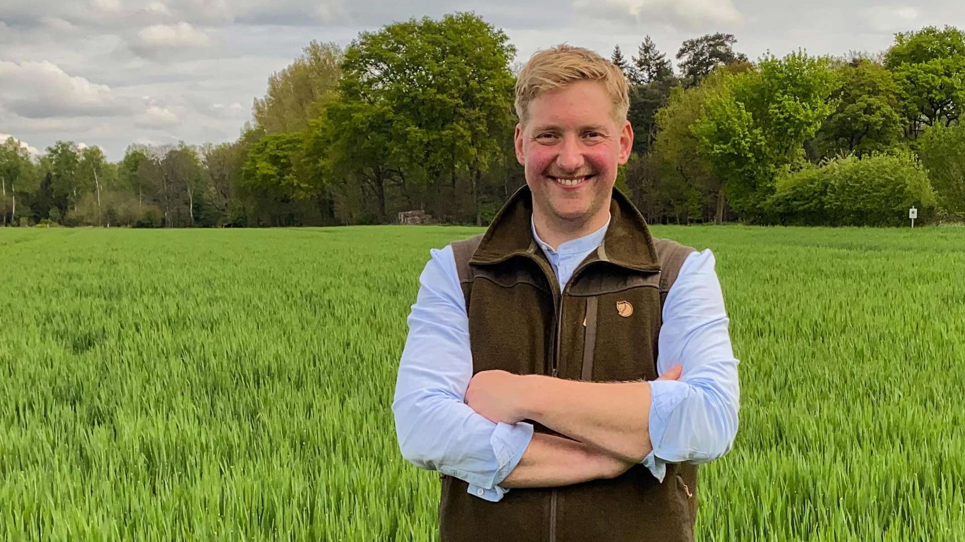 Bei Bauer sucht Frau 2025 steht Kandidat Daniel mit verschränkten Armen in einem grünen Getreidefeld. Mit Weste und Hemd blickt er freundlich in die Kamera vor bewaldetem Hintergrund.