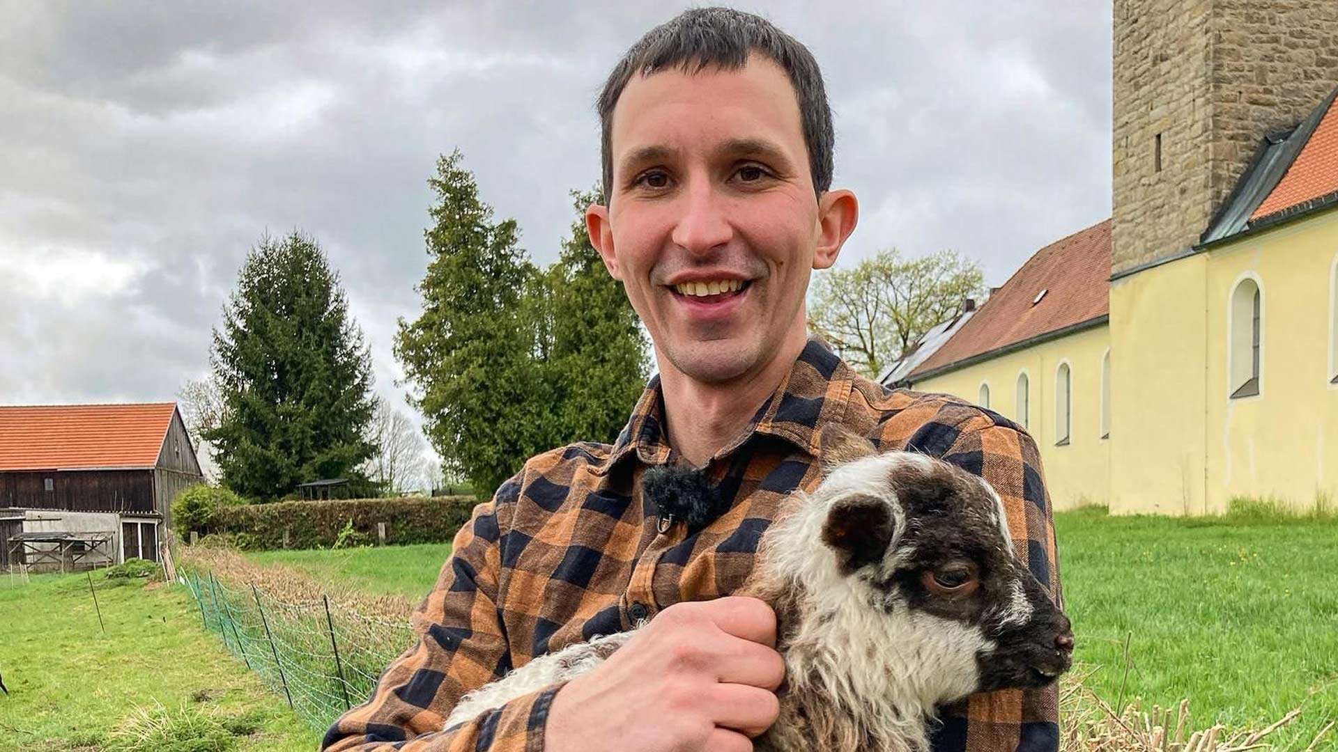 Bei Bauer sucht Frau 2025 hält Kandidat Michael ein schwarz-weißes Lämmchen im Arm. Mit kariertem Hemd steht er lächelnd auf einer grünen Wiese vor ländlicher Kulisse mit Kirche und Hof.