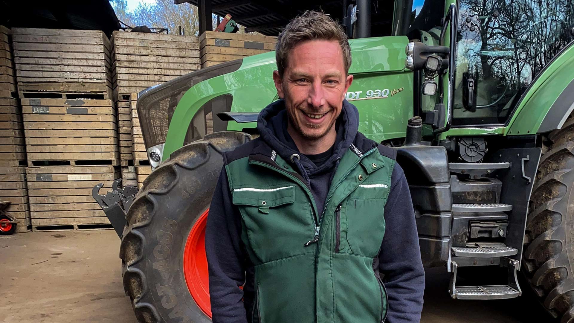 Bei Bauer sucht Frau 2025 steht Kandidat Andreas lächelnd vor einem grünen Traktor in einer Scheune. Er trägt eine grüne Arbeitsweste und vermittelt einen sympathischen, bodenständigen Eindruck auf seinem Hof.
