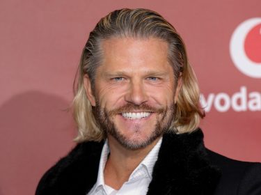 Auf dem Bild zur Übersicht Alle Bachelor ist Paul Janke vor einer roten Fotowand mit Vodafone-Logo zu sehen. Er trägt ein schwarzes Jackett mit Fellkragen und ein weißes Hemd. Mit einem strahlenden Lächeln und zurückgekämmten blonden Haaren wirkt er selbstbewusst und charmant – ganz im Stil des wohl bekanntesten Junggesellen der Sendung.