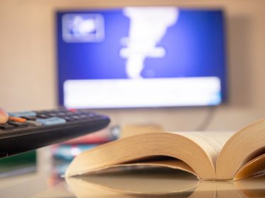 Ein aufgeschlagenes Buch liegt im Vordergrund auf einem Glastisch, während im Hintergrund unscharf ein Fernseher mit Nachrichtenbildschirm zu sehen ist. Eine Hand hält eine Fernbedienung in Richtung TV. Das Bild symbolisiert die Verbindung von Literatur und Fernsehen und steht stellvertretend für die kommenden Buchverfilmungen 2025, bei denen Romane zum Leben erweckt werden.